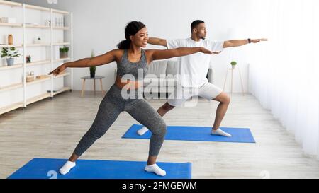 Strength yoga exercises. Fit black couple standing in warrior pose, exercising on sports mats at home, copy space