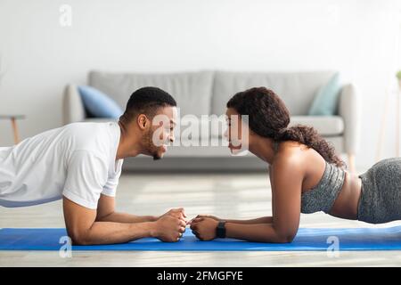 Side view of black couple standing in elbow plank, facing each other, having fun while doing domestic strength workout Stock Photo