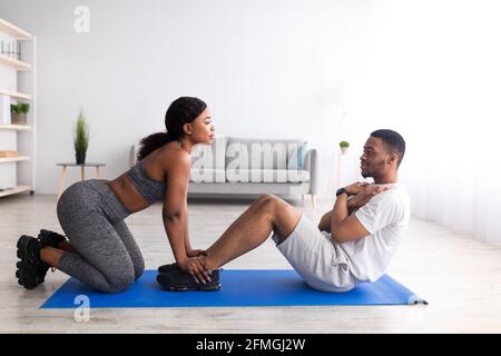 Millennial black guy doing abs exercises with his girlfriend, training as team at home, side view Stock Photo