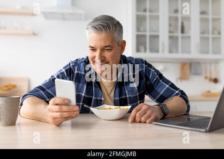Matured man using smartphone while having healthy breakfast in kitchen at home, sitting at table and eating cereals Stock Photo
