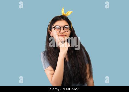 Let me think. Doubtful Indian teen girl touching her chin with finger, remembering something over blue studio background Stock Photo