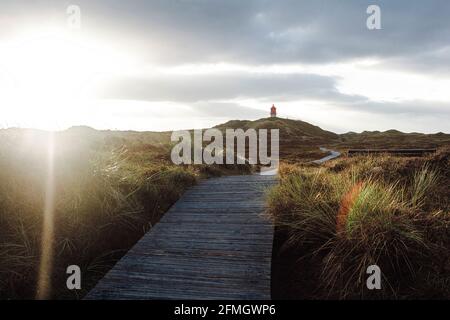 View against sun on summer landscape at North Sea coast. Boardwalk tourist pathway in nature leading to lighthouse in distance. Cloudy sky before suns Stock Photo