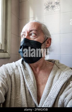 Portrait of an sad and worried old bearded man with protective mask in a bathrobe, looking out the bathroom window while there is a state of quarantin Stock Photo
