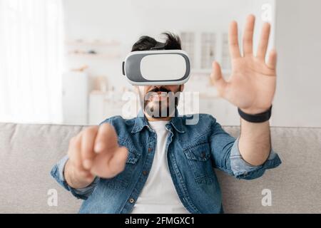 Future technology concept. Man playing video games wearing virtual reality glasses and gesturing, sitting on couch Stock Photo