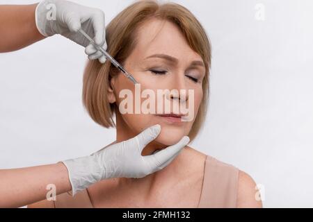 Attractive mature woman getting anti-aging procedure, receiving rejuvenating shot on light studio background Stock Photo