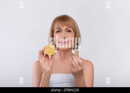 Fruit acids for skin care concept. Lovely mature woman holding lemon and rejuvenating serum on light studio background Stock Photo