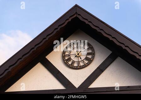 Large clock on a private house. Street clock in the bay window with roman numerals. Outdoor brown metal clock on white wooden roof. Stock Photo