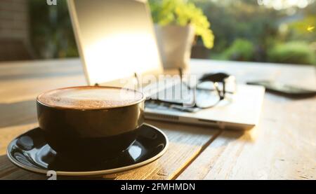Beautiful golden morning sunshine on an outdoor table setting with hot coffee, a laptop computer, mobile phone, glasses and mouse set up for online sh Stock Photo