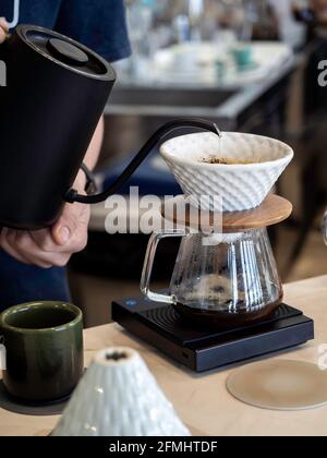 Drip coffee brewing in cafe. Barista pouring hot water from kettle into white ceramic coffee dripper on pot on electric hot stove on wooden table in c Stock Photo