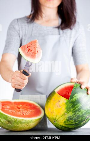A young caucasian woman is cutting a wedge out of a ripe watermelon using a knife. Melon is ripe with field spot. Light and airy summer season fruit c Stock Photo