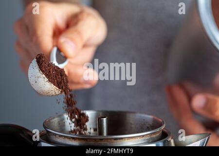 A caucasian woman is taking a teaspoon of ground coffee from a jar using a plastic measuring spoon and putting it into a percolator for brewing homema Stock Photo