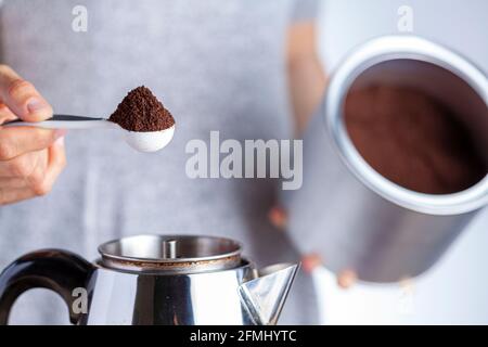 A caucasian woman is taking a teaspoon of ground coffee from a jar using a plastic measuring spoon and putting it into a percolator for brewing homema Stock Photo