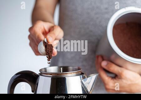 A caucasian woman is taking a teaspoon of ground coffee from a jar using a plastic measuring spoon and putting it into a percolator for brewing homema Stock Photo
