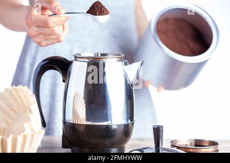 A caucasian woman is taking a teaspoon of ground coffee from a jar using a plastic measuring spoon and putting it into a percolator for brewing homema Stock Photo