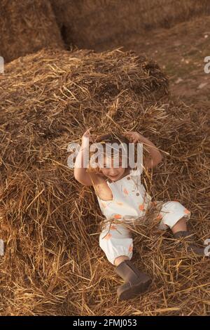 From above cheerful adorable child in overalls playing with hay lying down on straw bales in countryside Stock Photo