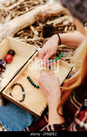 From above crop anonymous craftsWoman creating handmade bijouterie while using semiprecious stones and sitting on beach near sea Stock Photo