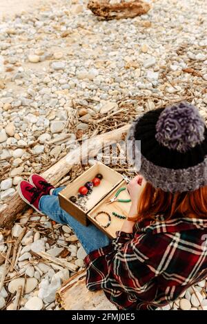 From above crop anonymous craftsWoman creating handmade bijouterie while using semiprecious stones and sitting on beach near sea Stock Photo