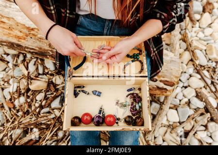 From above crop anonymous craftsWoman creating handmade bijouterie while using semiprecious stones and sitting on beach near sea Stock Photo