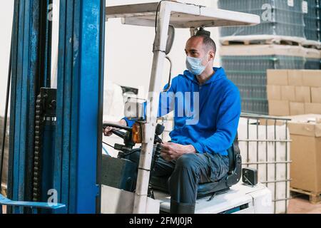 Male engineer in sterile mask driving vehicle and looking forward against cardboard boxes and bottles with beer in factory Stock Photo