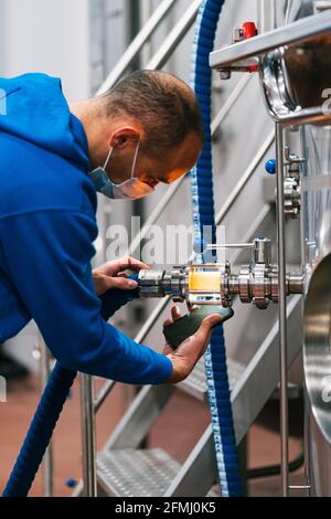 Side view of male worker in mask with cellphone checking up liquid in tube of lauter tun in beer factory during covid 19 pandemic Stock Photo