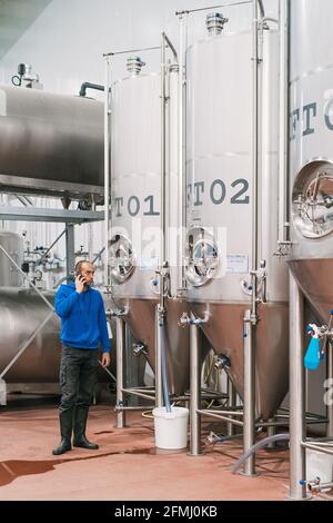 Side view of male entrepreneur talking on cellphone against stainless steel vessels on wet floor in beer factory Stock Photo