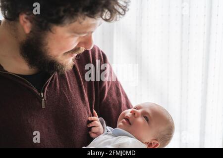 Adult bearded dad with curly hair cuddling cute little child looking at each other in house Stock Photo