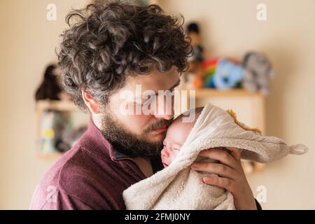Adult bearded dad with curly hair cuddling cute little child in soft blanket in house Stock Photo