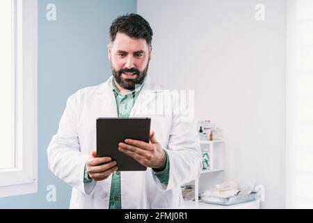 Mature beaded male medic in robe using tablet in clinic looking at screen Stock Photo