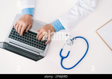 From above cropped unrecognizable male doctor in uniform typing on netbook at table with stethoscope in clinic Stock Photo