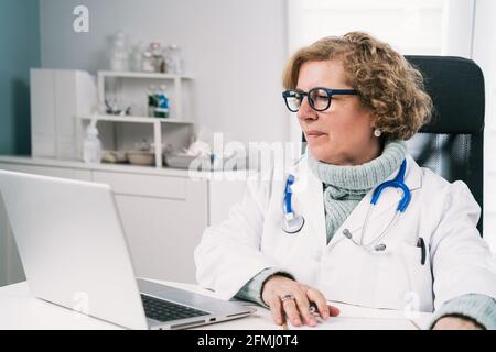 Mature female doctor in eyeglasses with stethoscope sitting at table with netbook while working in hospital Stock Photo