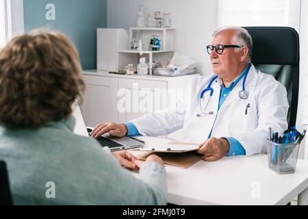 Elderly male doctor in uniform and eyeglasses typing on netbook at table against unrecognizable Woman during checkup in clinic Stock Photo