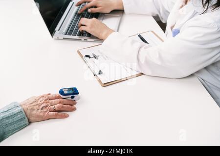 Crop unrecognizable doctor measuring blood oxygen of patient with heart rate monitor at table with stethoscope in clinic while using laptop Stock Photo