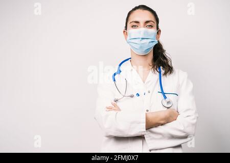 Self assured female doctor in uniform with stethoscope and sterile mask looking at camera with folded arms on white background during coronavirus pand Stock Photo