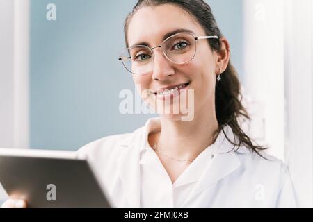 Young happy female medic in robe and eyeglasses using tablet in clinic looking at camera Stock Photo