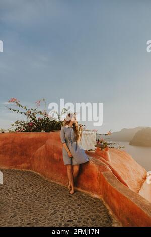 Full length young female in sundress sitting on coastal town embankment railing and admiring picturesque views on sunny day in Santorini Stock Photo