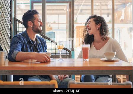 Cheerful ethnic couple looking at each other while having fun at wooden table with coffee and refreshing drinks Stock Photo