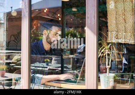 Through glass view of adult ethnic male remote employee working on netbook at table Stock Photo