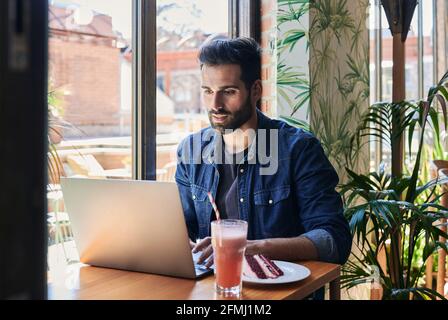 Side view of ethnic male employee working on netbook at table with tasty cake piece and drinks Stock Photo