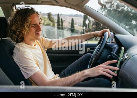 Side view of positive young male driver checking direction on GPS navigator on smartphone while traveling through countryside in summer day Stock Photo