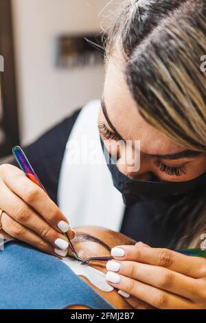 Cosmetologist with tweezers applying fake eyelashes for extension on eye of ethnic client with face protective mask in salon during coronavirus pandem Stock Photo