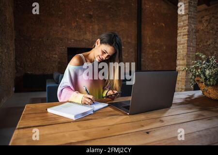 Content female remote employee with cellphone and netbook taking notes in notepad at table in house Stock Photo