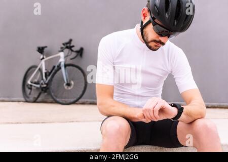 Male biker in protective helmet and sunglasses watching heart rate on wearable bracelet while sitting against bicycle on three color background Stock Photo