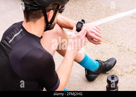 From above male biker in protective helmet and sunglasses watching heart rate on wearable bracelet while sitting on floor Stock Photo