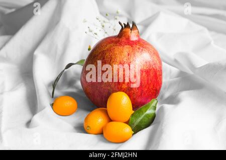 Bright whole fresh pomegranate with cumquats and leaves with sprigs on creased fabric on white background Stock Photo
