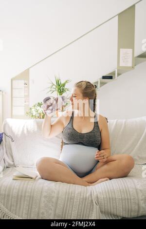 Young pregnant female in sports clothes with bottle of water sitting with crossed legs on sofa at home Stock Photo
