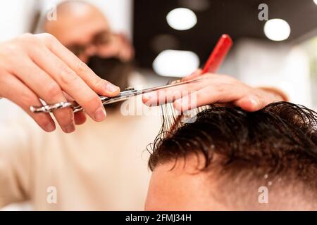 Male stylist trimming wet hair of crop anonymous client with scissors in hairdressing salon on blurred background Stock Photo