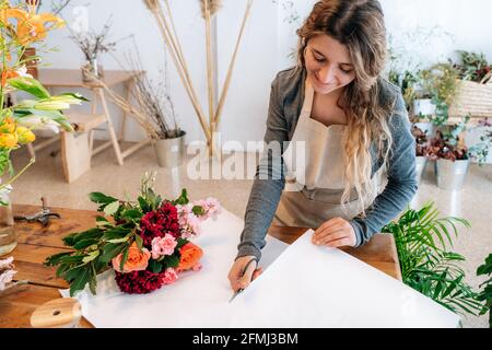 From above of crop happy ethnic young female florist in apron smiling and cutting wrapping paper while arranging elegant bouquet of fresh assorted col Stock Photo