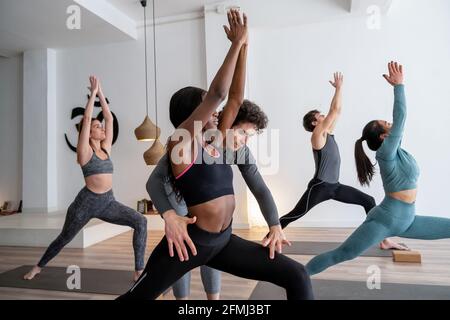 Side view of company of diverse people practicing yoga in Warrior pose with help of trainer in studio Stock Photo