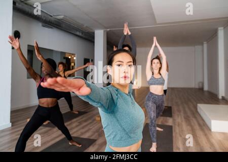 Focused Asian female standing in Virabhadrasana and practicing mindfulness during yoga lesson with diverse people in studio Stock Photo