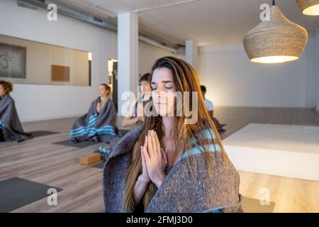 Young female wrapped in blanket meditating with Namaste hands in studio with diverse people practicing yoga together Stock Photo